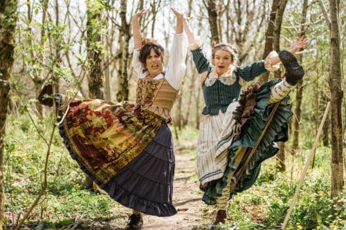 Theatre artists The Grimm Sisters in the woods in a playful pose with one foot above the ground and hands up in the air
